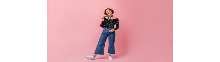 Women in long sleeve polka dot top, wide leg capri pants, and white canvas sneakers with a hat in front of a pink background